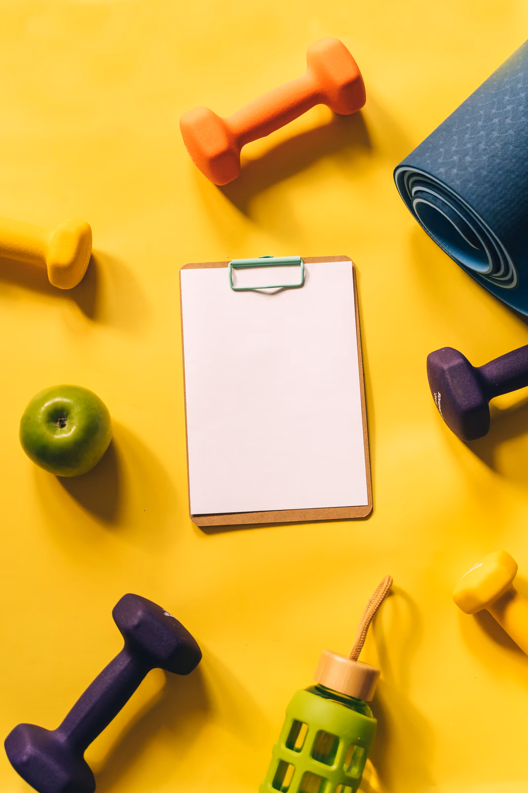 Dumbbells, a yoga mat, a green apple, a water bottle and a clipboard on a yellow background